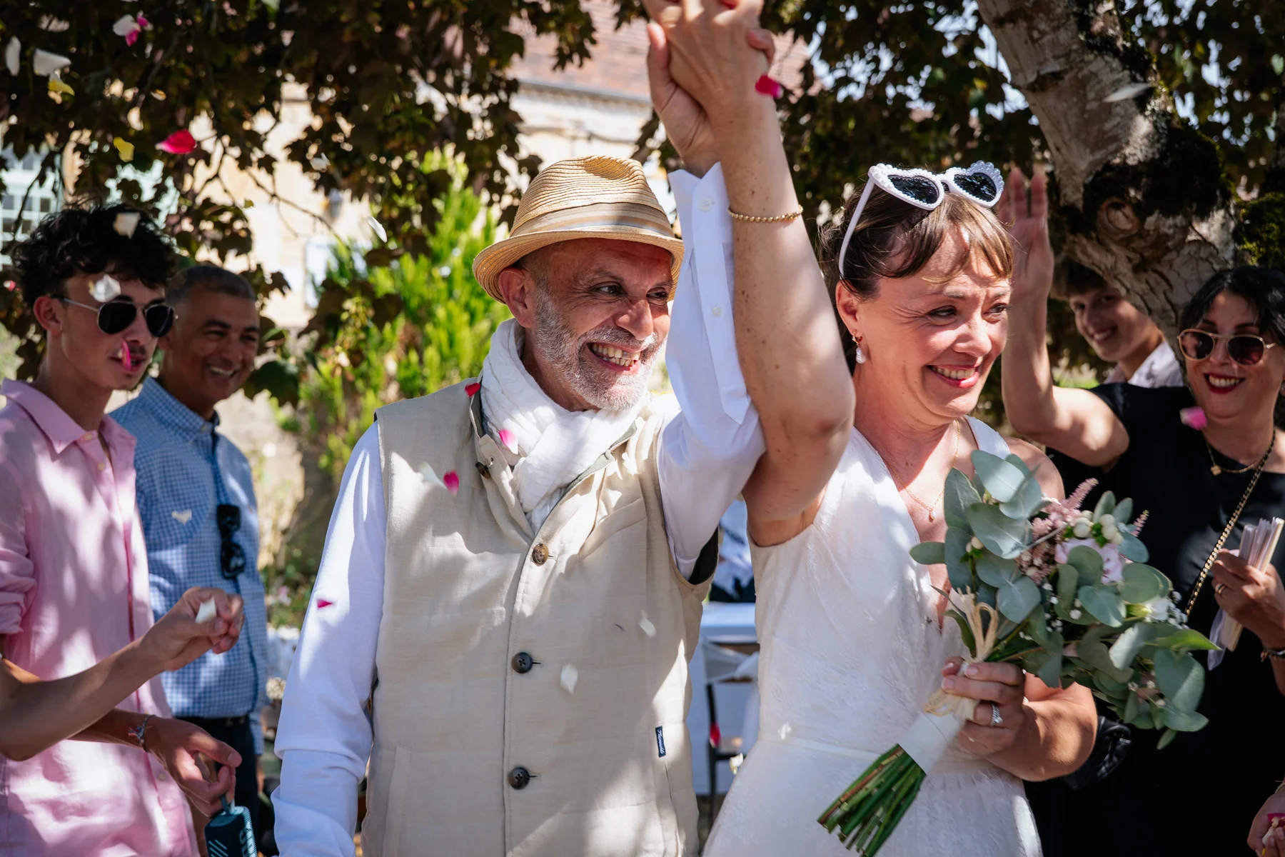 Des mariés sourient, les bras levés sous une pluie de pétales, entourés de leurs invités.