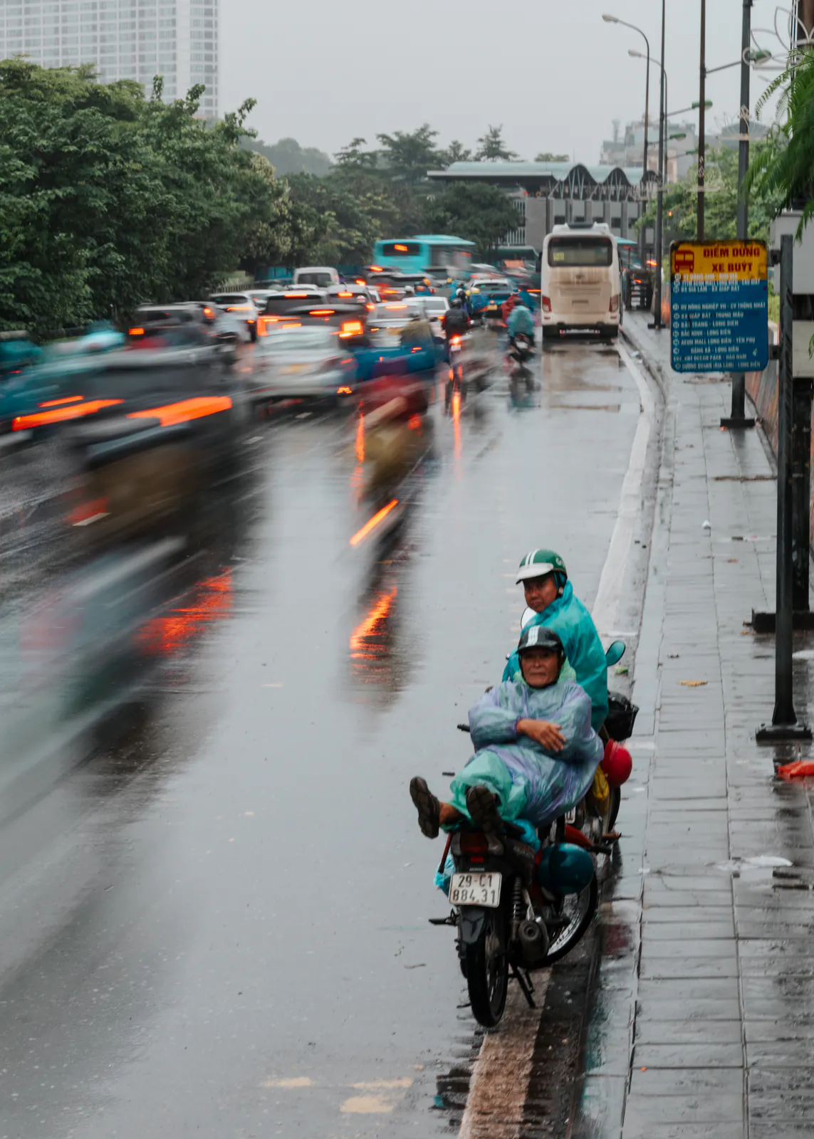 Des scooters traversent une avenue sous la pluie, entre flou de mouvement et reflets orange.