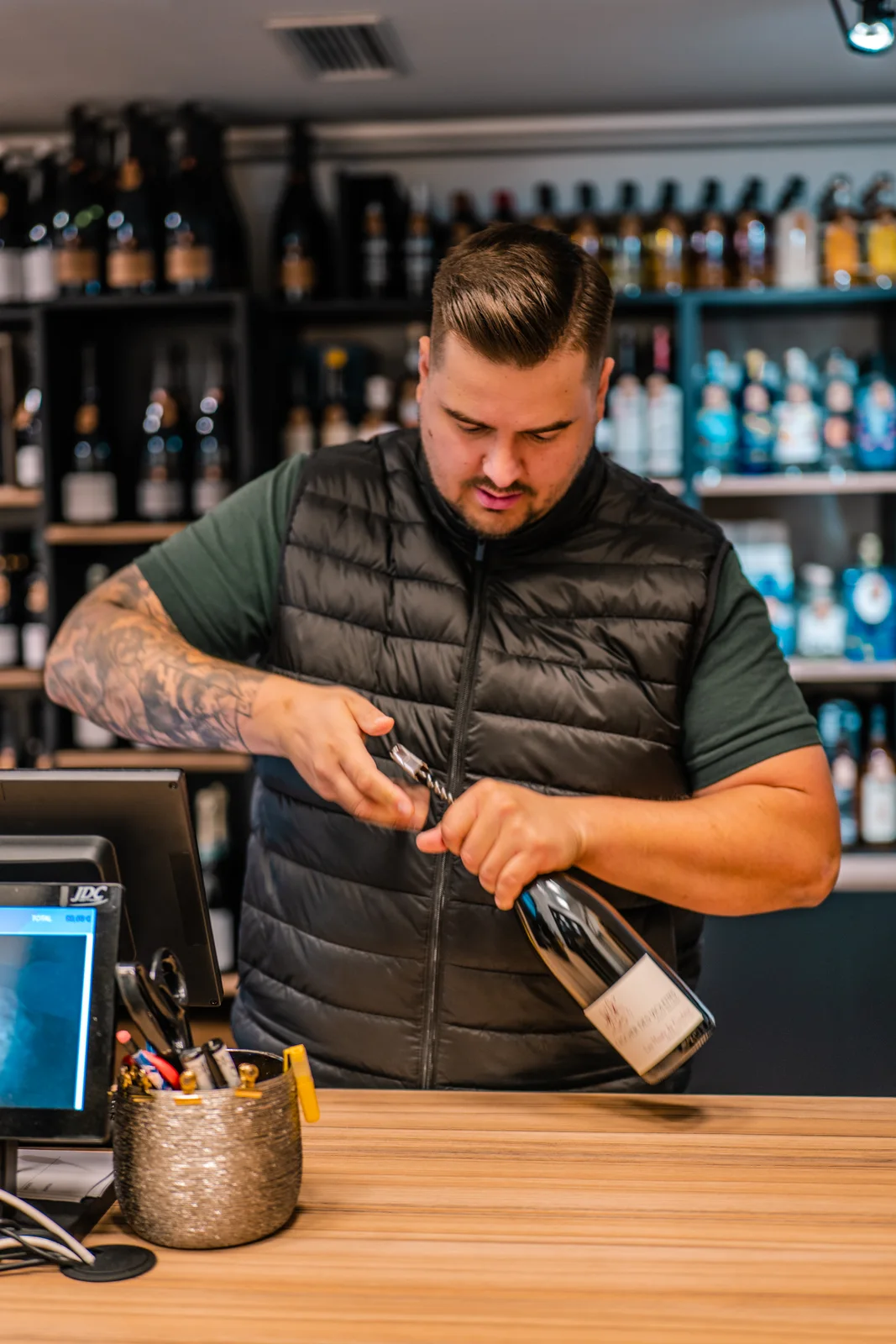 Un caviste ouvre une bouteille derrière son comptoir, entouré de ses références.