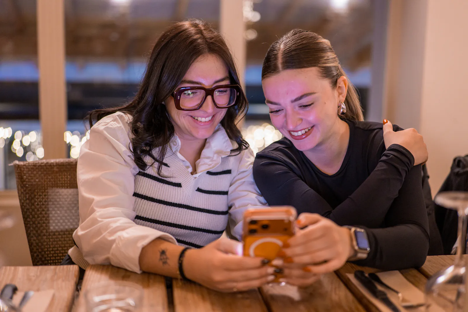 Deux femmes rient ensemble en regardant un téléphone à une table.