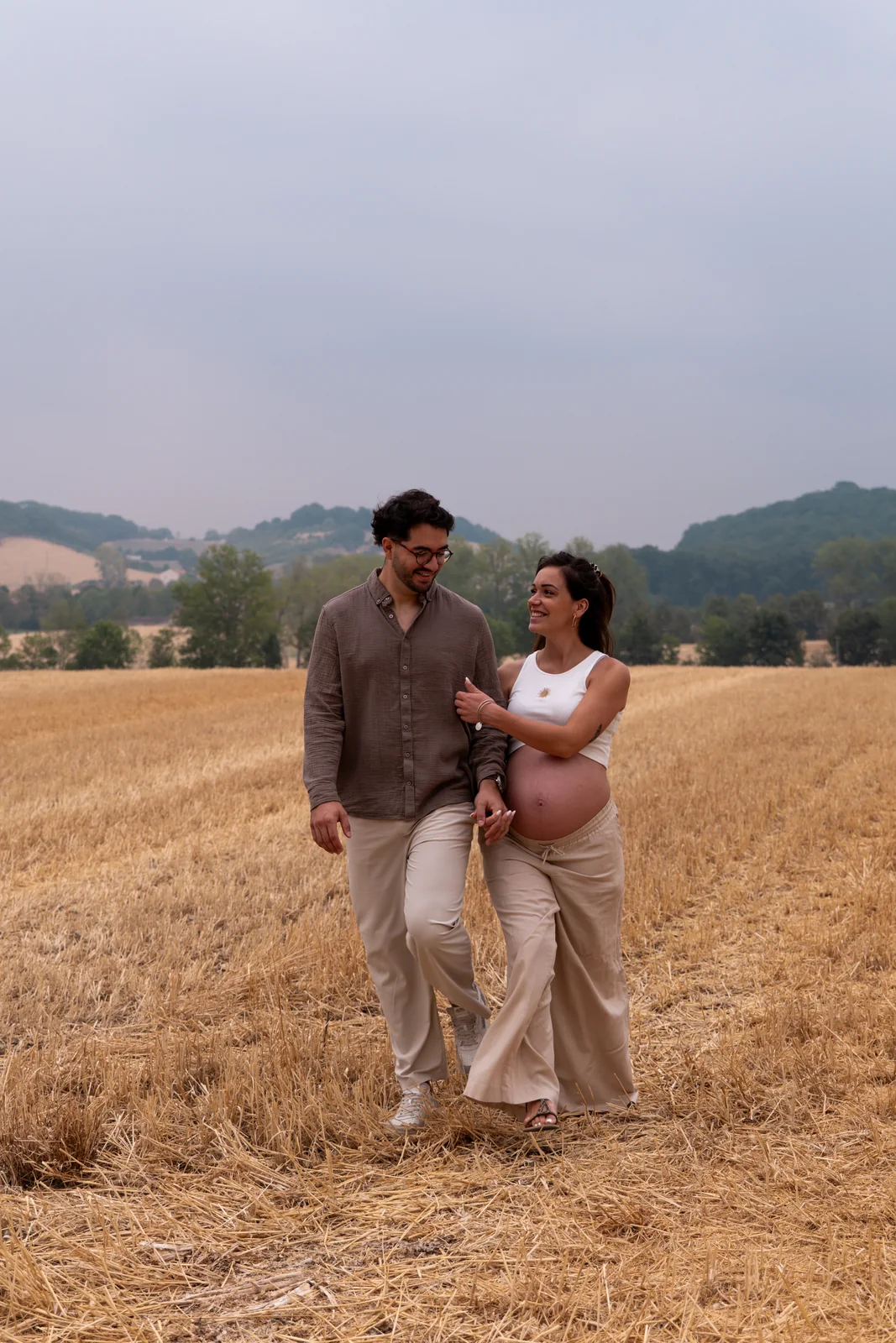 Les futurs parents marchent enlacés dans les blés, complices et détendus.