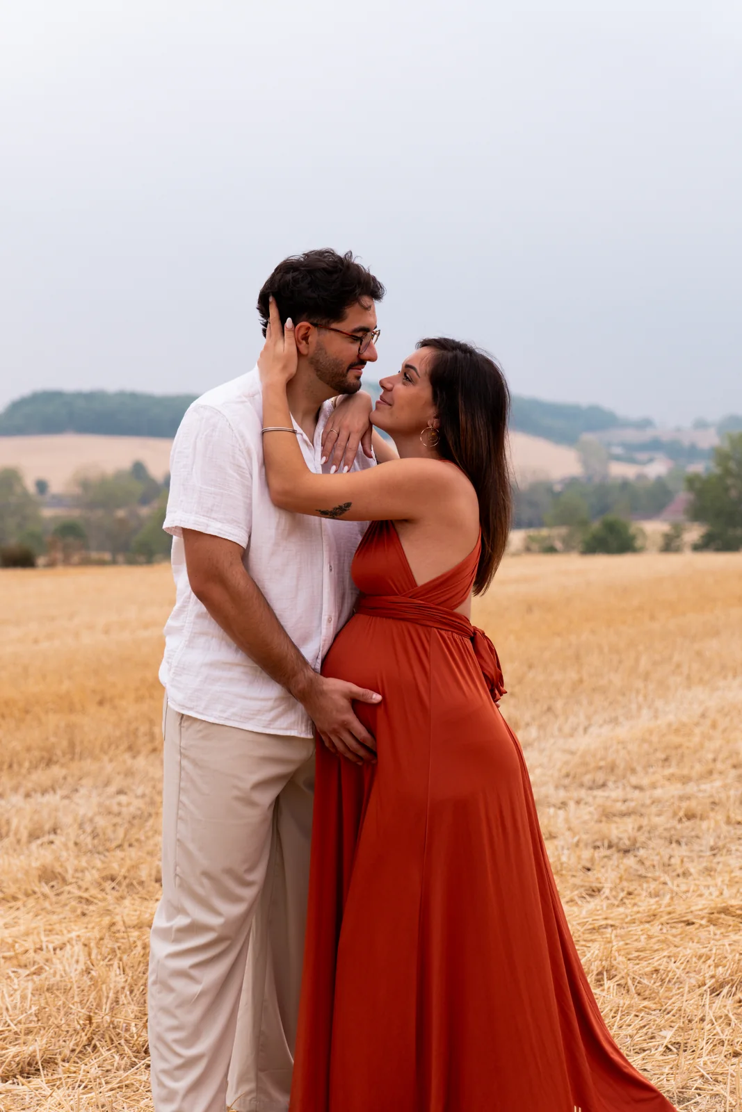 Les futurs parents s’embrassent dans un champ, le ventre rond au centre de leur étreinte.