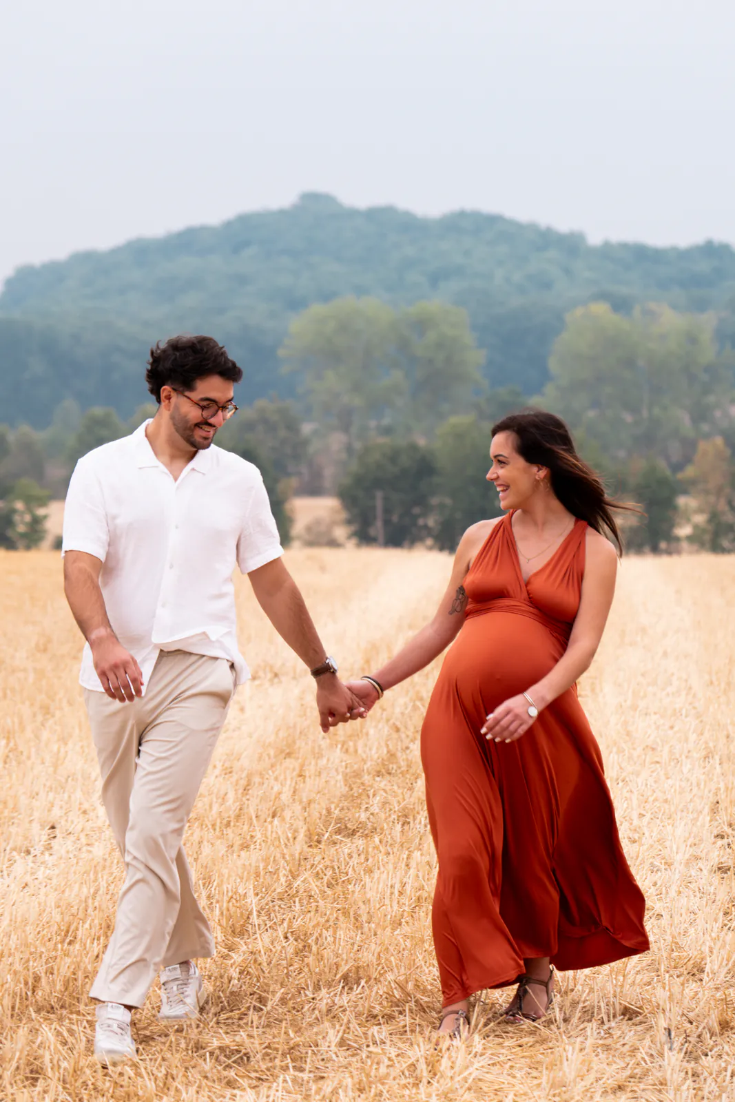Un couple attend un enfant et marche main dans la main dans un champ, la future mère vêtue de rouge.