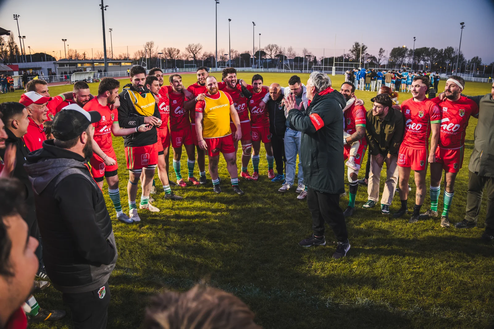 Une équipe de rugby réunie en cercle écoute les mots d’après-match au coucher du soleil.