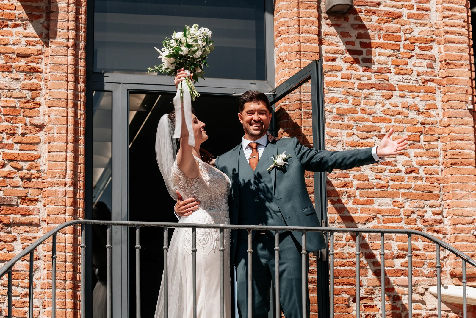 Deux jeunes mariés lèvent les bras avec joie à la sortie de la cérémonie, devant une façade de briques.