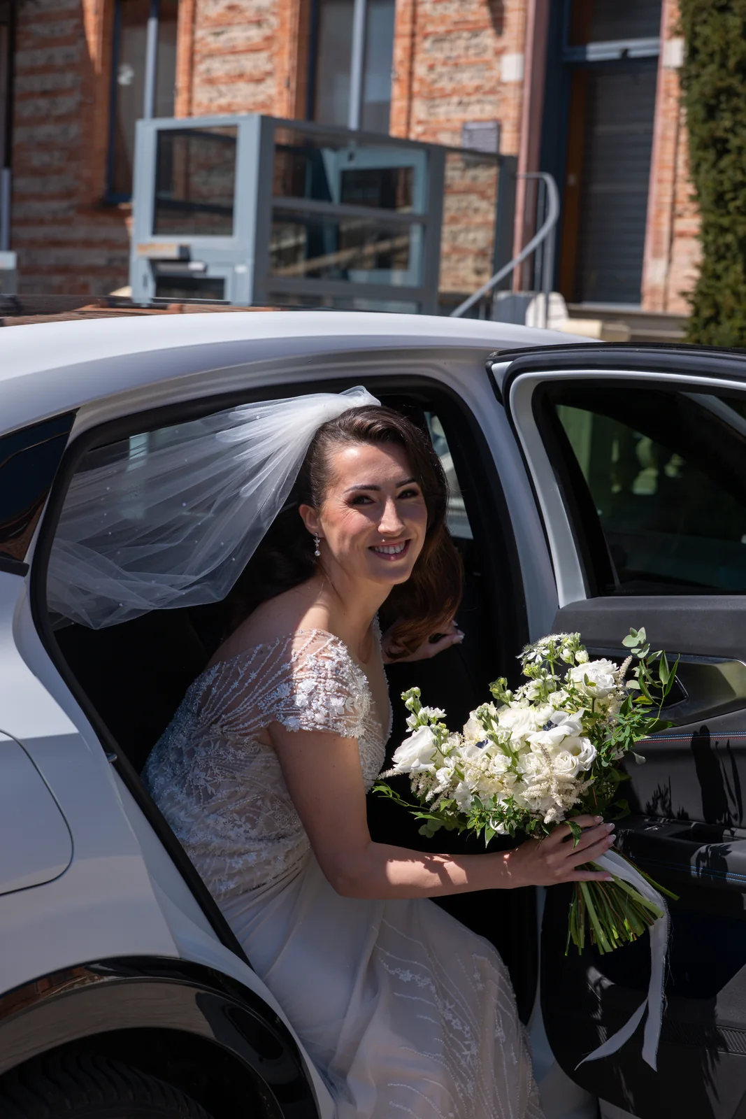 Une mariée sourit en descendant de voiture, son bouquet blanc à la main.