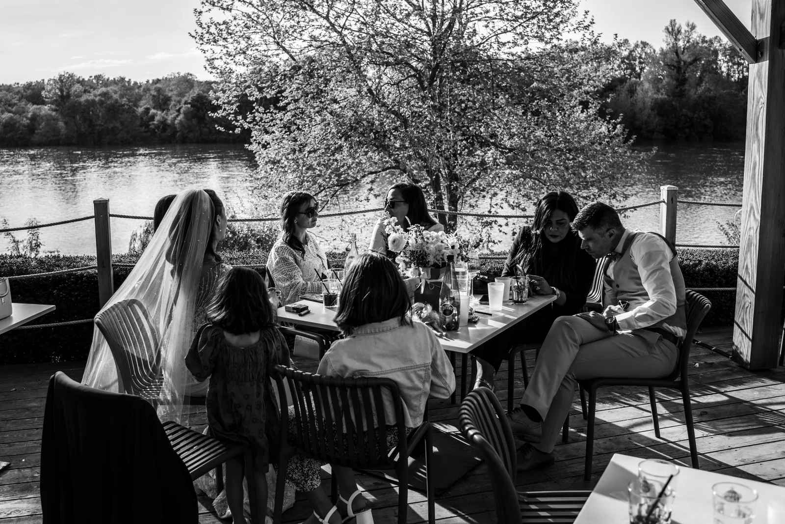 Les mariés et leurs proches partagent une longue table au bord de l’eau, en noir et blanc.